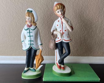 May include: Two vintage porcelain figurines of girls wearing white coats with red and green accents. One girl is holding an umbrella and the other is holding a brown bag. The figurines are standing on green bases.