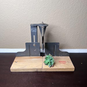 Puede incluir: Una caja de ingletes Stanley vintage con base de madera y componentes metálicos grises. La guía de sierra está colocada sobre la base de madera. Una pequeña suculenta verde se coloca en la base de madera. El texto "STANLEY MADE IN U.S.A." es visible.