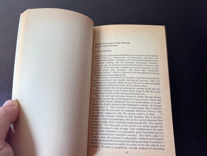 May include: A page from a book titled "Social Functions of the Prisons in the United States" by Bettina Aptheker. The text discusses the use of euphemisms like "correctional facilities" and "inmates" to avoid acknowledging the reality of prisons and the existence of political prisoners in the United States.
