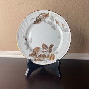 May include: A white porcelain plate with a gold rim and a floral design in gold and gray. The plate is on a black stand.