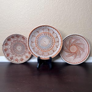 May include: Three ceramic plates with intricate white designs on a brown background. The plates have a raised rim and are displayed on a dark wooden surface.