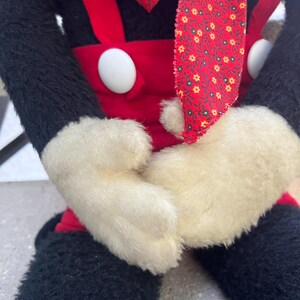 May include: A plush doll featuring a black body, red overalls with white buttons, and white gloves. The doll has a red patterned necktie and is seated. The doll's arms are crossed in front of its body.