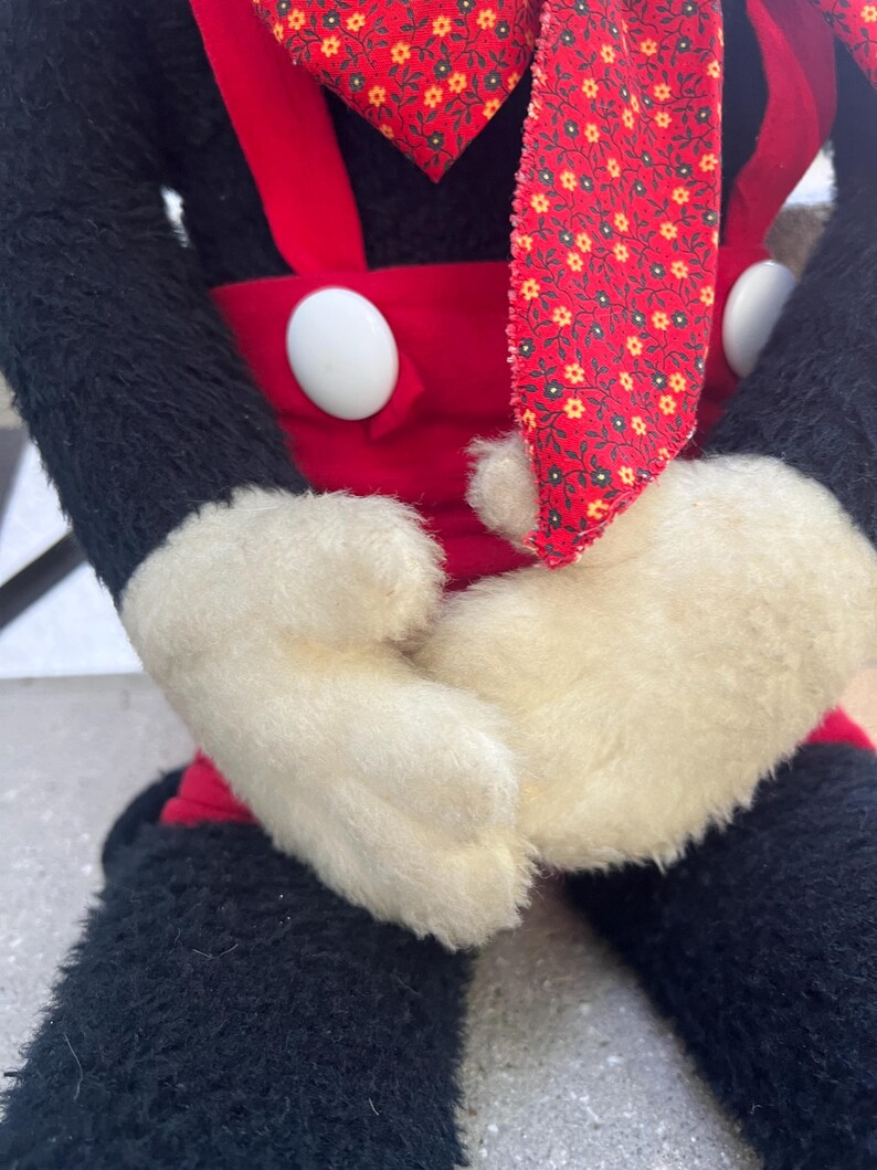 May include: A plush doll featuring a black body, red overalls with white buttons, and white gloves. The doll has a red patterned necktie and is seated. The doll's arms are crossed in front of its body.