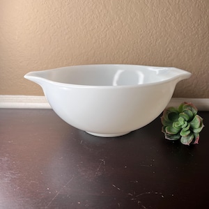 Pode incluir: Uma tigela de mistura de cerâmica branca com duas alças. A tigela está sobre uma superfície de madeira marrom escura.