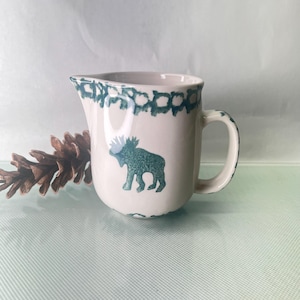 Puede incluir: Cremera de cerámica color crema con una silueta de alce turquesa y borde decorativo. El asa y el borde presentan detalles turquesa. Una piña descansa junto a la cremera, sugiriendo un tema rústico.