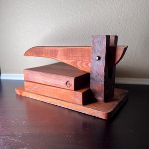 May include: A wooden cigar press with a lever and a base made of stacked wooden planks. The press is made of dark reddish-brown wood.