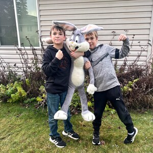 May include: Two boys pose with a large plush Bugs Bunny toy. The plush rabbit is gray and white with long ears and a carrot in its mouth. The boys are wearing casual clothing, standing on green grass.