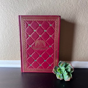 May include: A red leather-bound book with a gold-coloured border and a decorative pattern of green leaves. The book has a gold-coloured crest on the front cover.