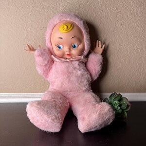 May include: A vintage baby doll with a pink plush body and hood. The doll has a plastic face with blue eyes, a red mouth, and a yellow swirl on top of its head. The doll is sitting with its arms outstretched.