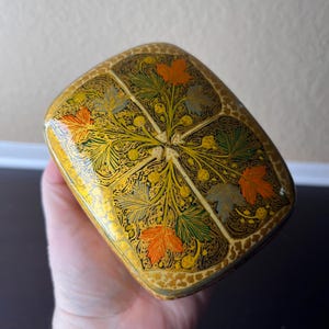 May include: A decorative, rectangular box with a rounded top. The box is painted gold with a floral design featuring orange, green, and gray leaves. The box is held in a hand.