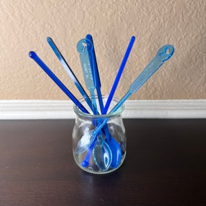 May include: A glass jar filled with blue plastic stirring spoons. The spoons have a variety of shapes and sizes, including a spoon with the text "The Lobster" printed on it.