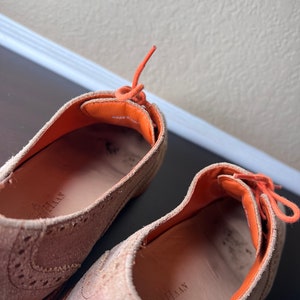 Puede incluir: Un par de zapatos de ante beige con cordones naranjas. Los zapatos tienen un dise&ntilde;o de brogue y el texto "MADE IN ITALY" es visible en el interior del zapato.