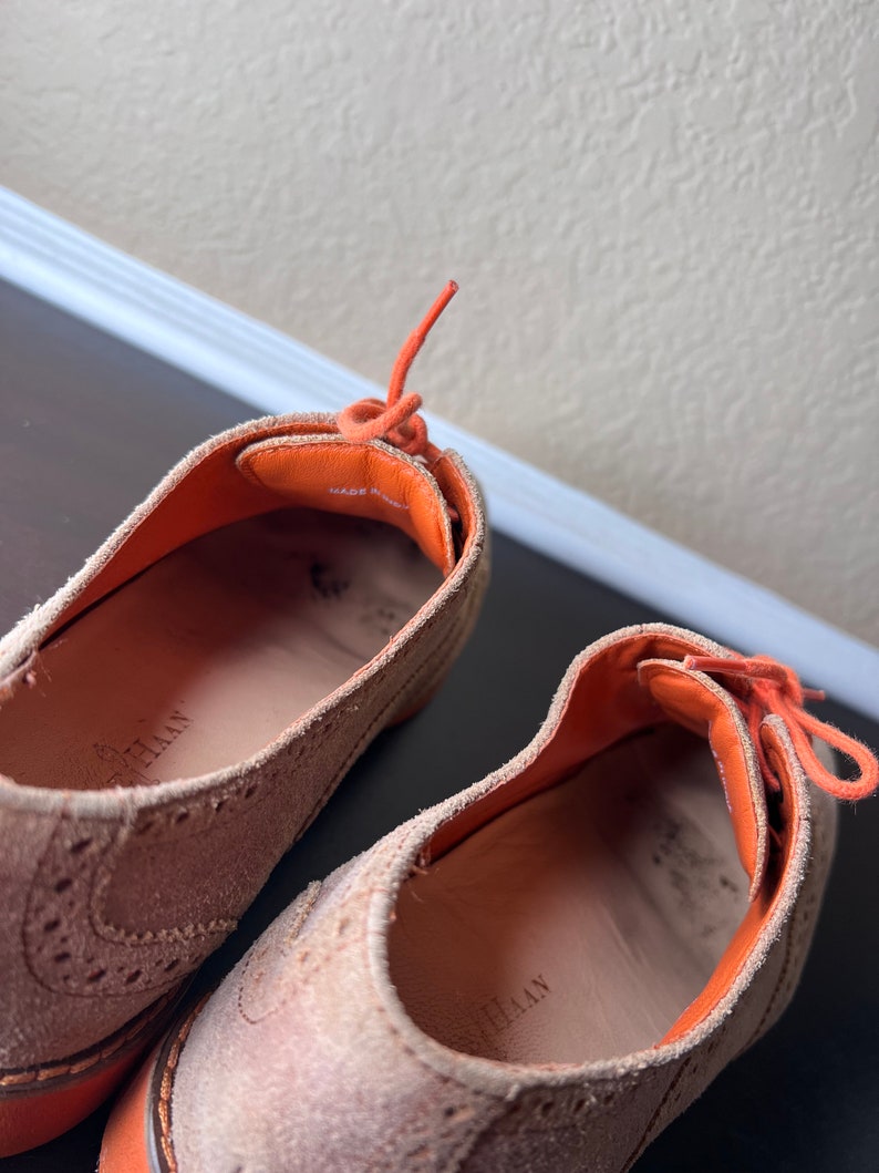 Puede incluir: Un par de zapatos de ante beige con cordones naranjas. Los zapatos tienen un dise&ntilde;o de brogue y el texto "MADE IN ITALY" es visible en el interior del zapato.