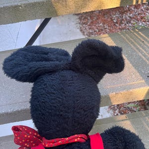 May include: A black plush toy with large, floppy ears and a red bow tie. The toy has red suspenders and a soft, textured appearance. The image is taken from a slightly elevated angle, showcasing the back of the toy.