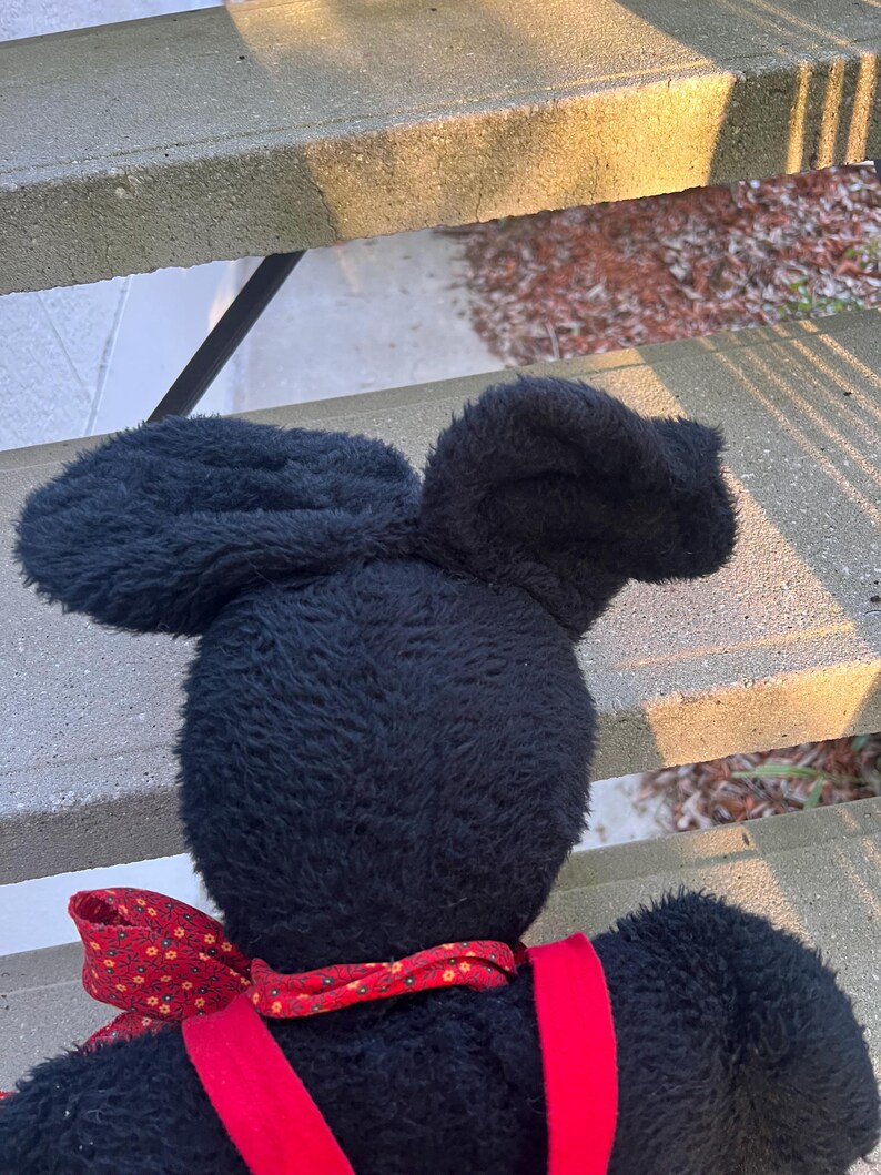 May include: A black plush toy with large, floppy ears and a red bow tie. The toy has red suspenders and a soft, textured appearance. The image is taken from a slightly elevated angle, showcasing the back of the toy.