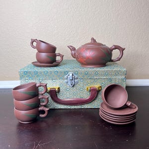 Puede incluir: Un juego de té de cerámica marrón con una tetera, tazas y platillos, todo en una caja de madera con un forro estampado en verde y dorado.