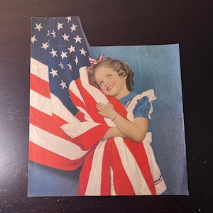 以下が含まれることがあります： 大きなアメリカ国旗を抱きしめる少女のヴィンテージ画像。彼女は白いトリムと赤いリボンが付いた青いドレスを着ています。旗の赤と白のストライプと星のある青いフィールドが目立っています。