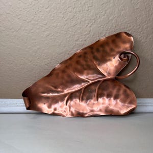 May include: Copper leaf-shaped decorative tray with a curled handle. The tray has a natural leaf vein pattern and a rich, reddish-brown color. The tray is made of copper and has a smooth, polished finish. A unique accent piece for home decor.