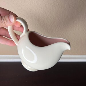 May include: Cream-colored ceramic creamer with a pink interior and a curved handle. The creamer has a rounded body and a spout for easy pouring. A classic piece for serving cream or milk.