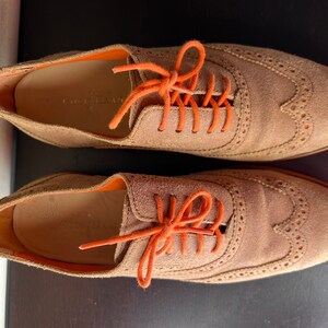 Puede incluir: Un par de zapatos oxford de ante marr&oacute;n con cordones naranjas. Los zapatos tienen un dise&ntilde;o de punta de ala y est&aacute;n atados con un lazo.