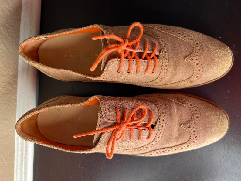 Puede incluir: Un par de zapatos oxford de ante marr&oacute;n con cordones naranjas. Los zapatos tienen un dise&ntilde;o de punta de ala y est&aacute;n atados con un lazo.