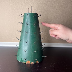 May include: A green, cone-shaped wooden object with numerous nails protruding from its surface. The object is placed on a dark surface. A finger is pointing at the object.