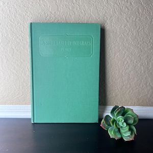 Könnte beinhalten: Ein türkisfarbenes Buch mit dem Titel "A Short Table of Integrals" von Peirce. Das Buch steht aufrecht und hat ein rechteckiges Etikett mit dem geprägten Titel auf dem Einband. Rechts vom Buch befindet sich eine grüne Sukkulentenpflanze mit rot gefärbten Blättern.