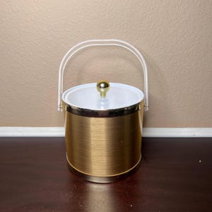 May include: A gold-colored ice bucket with a white lid and a clear plastic handle. The bucket has a gold knob on top of the lid and a gold band around the top and bottom. The bucket is cylindrical in shape.