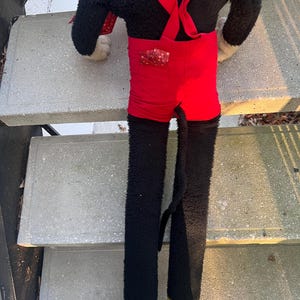 May include: A vintage plush toy featuring a black body, red shorts with suspenders, and yellow feet. The toy has a long tail and is positioned on concrete steps. The plush toy is a classic character.