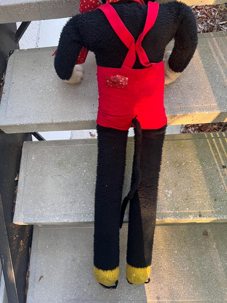 May include: A vintage plush toy featuring a black body, red shorts with suspenders, and yellow feet. The toy has a long tail and is positioned on concrete steps. The plush toy is a classic character.