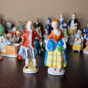 May include: Two porcelain figurines of a man and woman in traditional clothing. The man is wearing a red jacket with gold buttons, white pants with blue and red stripes, and a blue and green hat. The woman is wearing a blue and yellow dress with a green sash. Both figurines are standing on a white base.