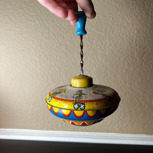 May include: A vintage toy top with a blue handle and a yellow, red, and blue painted design. The top features a circus scene with a clown, a horse, and a wagon.