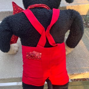May include: A plush black dog toy wearing red shorts with suspenders and a red bow tie. The shorts have a small pocket with a patterned fabric. The dog has white paws and a red collar.