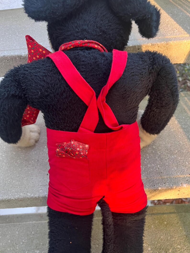 May include: A plush black dog toy wearing red shorts with suspenders and a red bow tie. The shorts have a small pocket with a patterned fabric. The dog has white paws and a red collar.