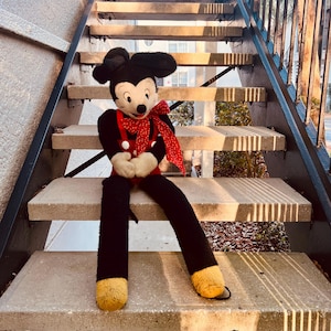 May include: A vintage Mickey Mouse plush toy with long legs, wearing a red outfit with white buttons and a red polka-dot bow tie. The plush is black, red, white, and yellow, and is sitting on concrete steps.