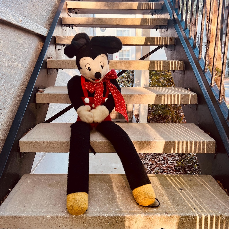 May include: A vintage Mickey Mouse plush toy with long legs, wearing a red outfit with white buttons and a red polka-dot bow tie. The plush is black, red, white, and yellow, and is sitting on concrete steps.