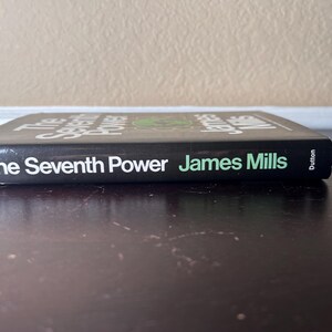 May include: A hardcover book titled "The Seventh Power" by James Mills. The book has a black cover with white text and a green graphic. The author's name is in green. The book is on a dark surface.