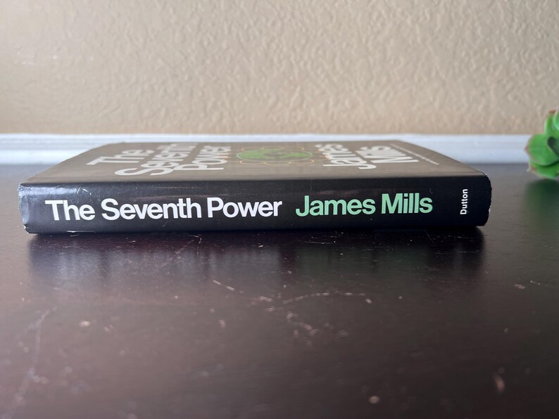 May include: A hardcover book titled "The Seventh Power" by James Mills. The book has a black cover with white text and a green graphic. The author's name is in green. The book is on a dark surface.