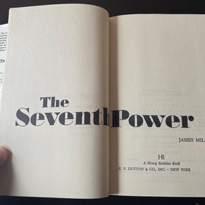 May include: Open book with the title "The Seventh Power" by James Mills. The book's cover is a light beige color with black text. The publisher is E.P. Dutton & Co., Inc., New York.