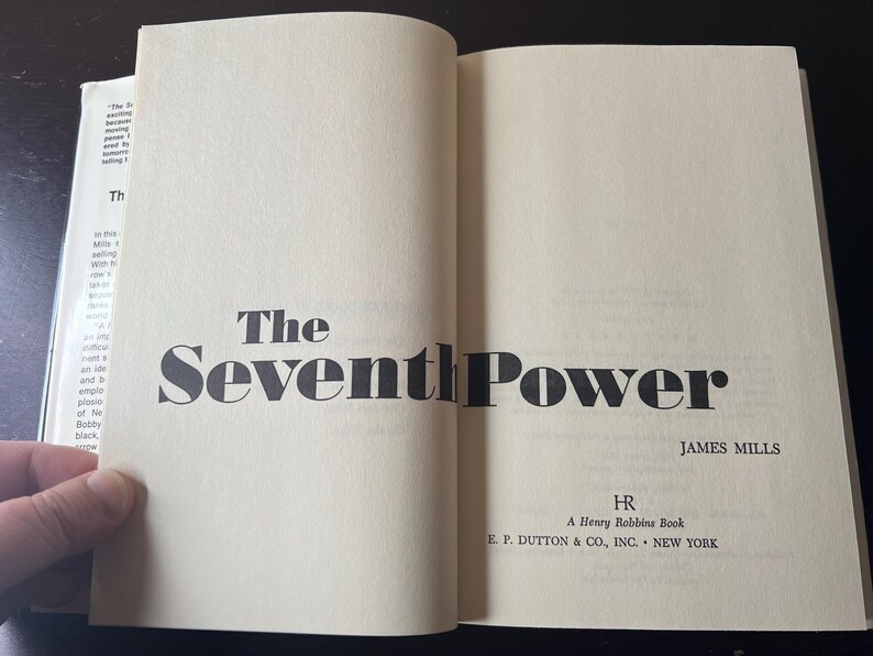 May include: Open book with the title "The Seventh Power" by James Mills. The book's cover is a light beige color with black text. The publisher is E.P. Dutton & Co., Inc., New York.