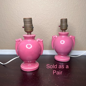 May include: A pair of pink ceramic lamp bases with a rounded body, a fluted neck, and two side handles. Each lamp features a metallic socket at the top and a white electrical cord extending from the base. The text Sold as a Pair is visible.