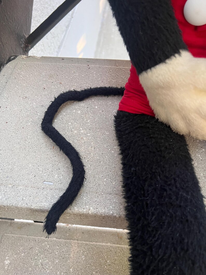 May include: A plush toy featuring a black and white furry arm and a black tail. The arm has a red sleeve and a white cuff. The tail is long and curved, lying on a concrete surface. The toy is likely a character from a cartoon.