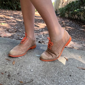 Puede incluir: Un par de zapatos Oxford de ante marr&oacute;n con cordones y suelas naranjas. Los zapatos tienen un dise&ntilde;o de punta de ala y se llevan puestos.