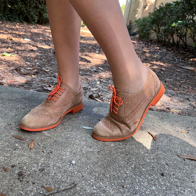 Puede incluir: Un par de zapatos Oxford de ante marr&oacute;n con cordones y suelas naranjas. Los zapatos tienen un dise&ntilde;o de punta de ala y se llevan puestos.
