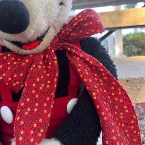 May include: A plush toy featuring a black and white face with a red bow and red overalls. The bow has a floral pattern with yellow and white flowers. The overalls have large white buttons. The toy's arms are black.