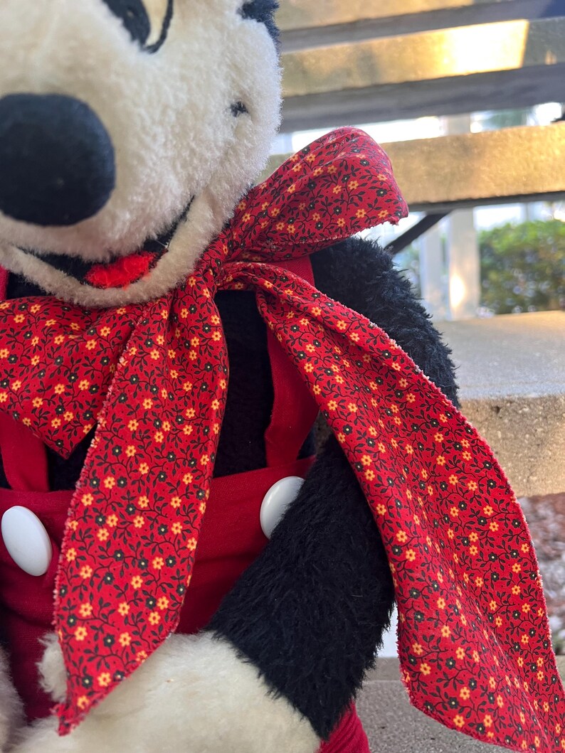 May include: A plush toy featuring a black and white face with a red bow and red overalls. The bow has a floral pattern with yellow and white flowers. The overalls have large white buttons. The toy's arms are black.
