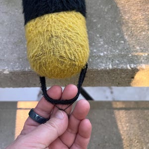 May include: A plush toy with a black and yellow color scheme. The toy is cylindrical, with a black top and a yellow bottom. A black loop is attached to the bottom.