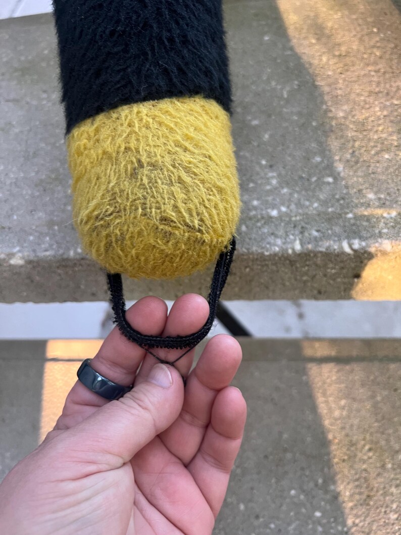 May include: A plush toy with a black and yellow color scheme. The toy is cylindrical, with a black top and a yellow bottom. A black loop is attached to the bottom.