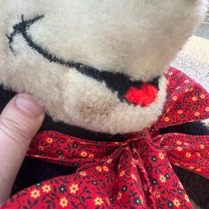 May include: Close-up of a vintage stuffed animal with a cream-colored face, black stitched details, and a red bow. The bow features a red background with a floral pattern. The toy's fur is soft and slightly worn.
