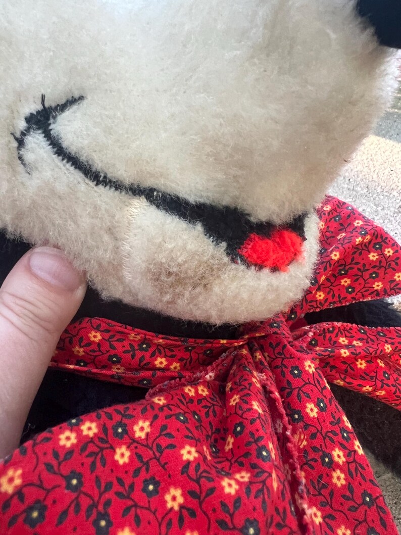 May include: Close-up of a vintage stuffed animal with a cream-colored face, black stitched details, and a red bow. The bow features a red background with a floral pattern. The toy's fur is soft and slightly worn.